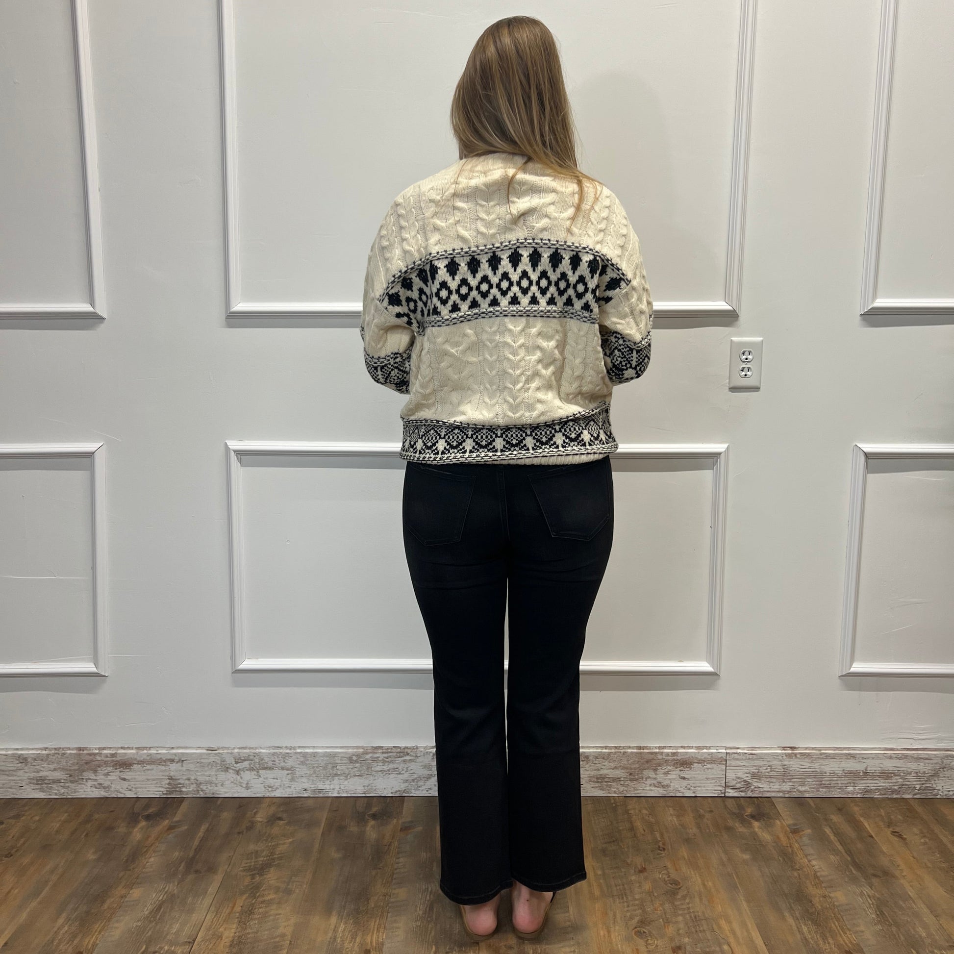 A woman stands with her back to the camera wearing a cream-colored cropped jacket with a black geometric pattern across the shoulders and sleeves, paired with black pants, in front of a white paneled wall.
