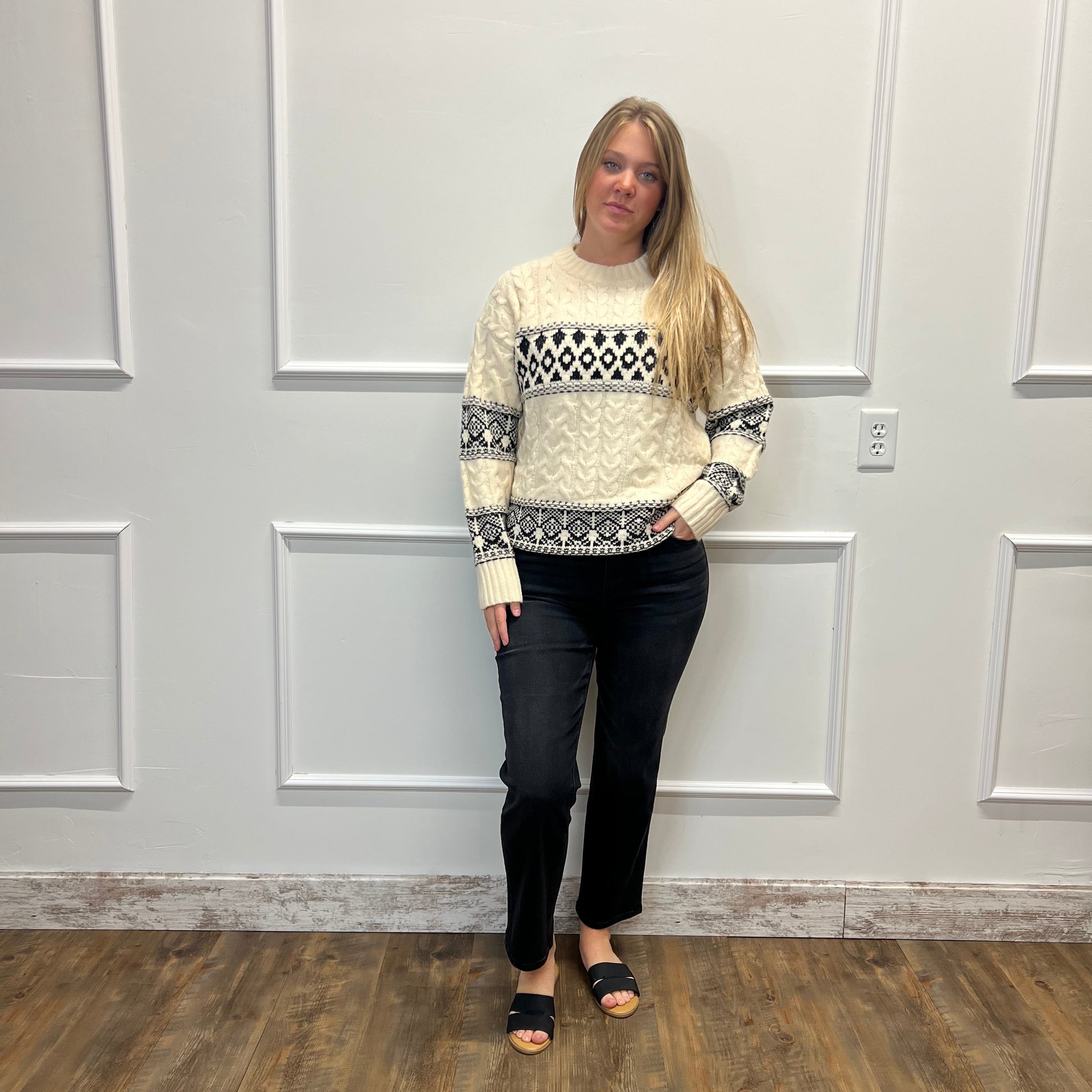 A woman stands indoors against a white paneled wall wearing a cream knit sweater with black geometric and floral patterns, black pants, and black slide sandals.