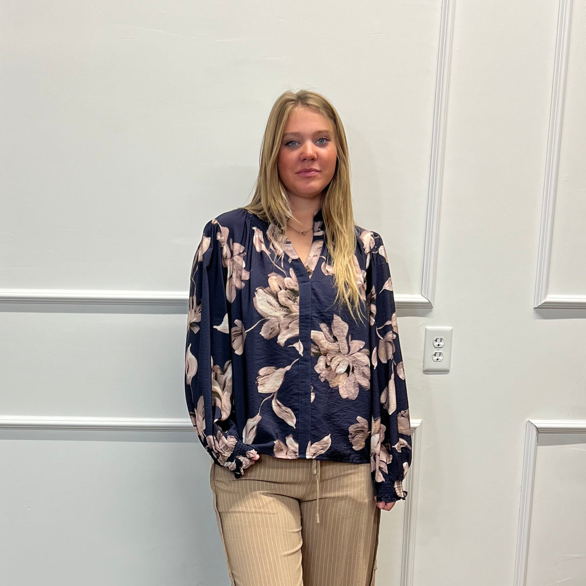 A woman stands in a room with white paneled walls and a wood floor, wearing a navy blue floral long-sleeve blouse and beige wide-leg pants.