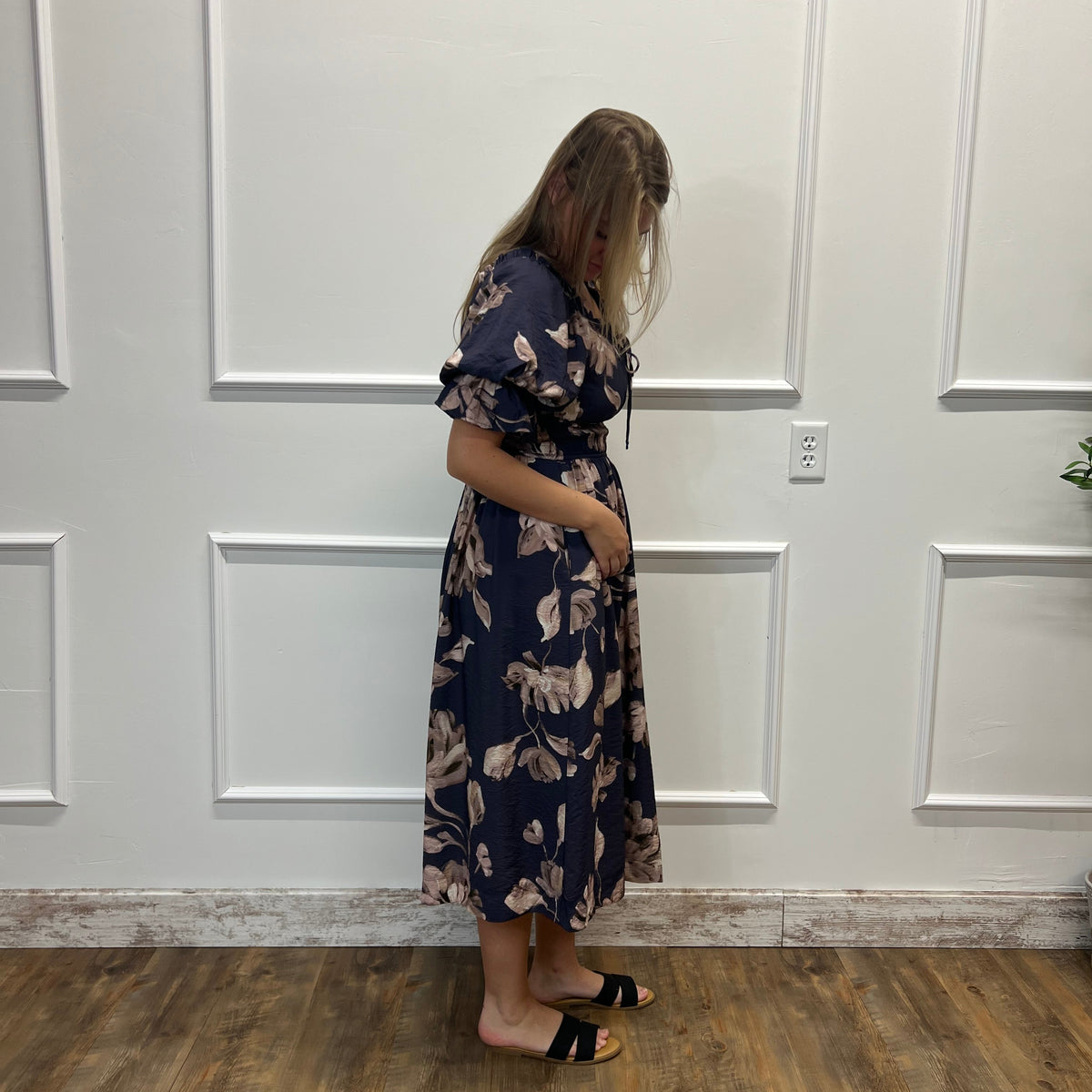 A woman wearing a navy blue midi dress with a beige floral pattern stands in profile against a white paneled wall on a wood floor.