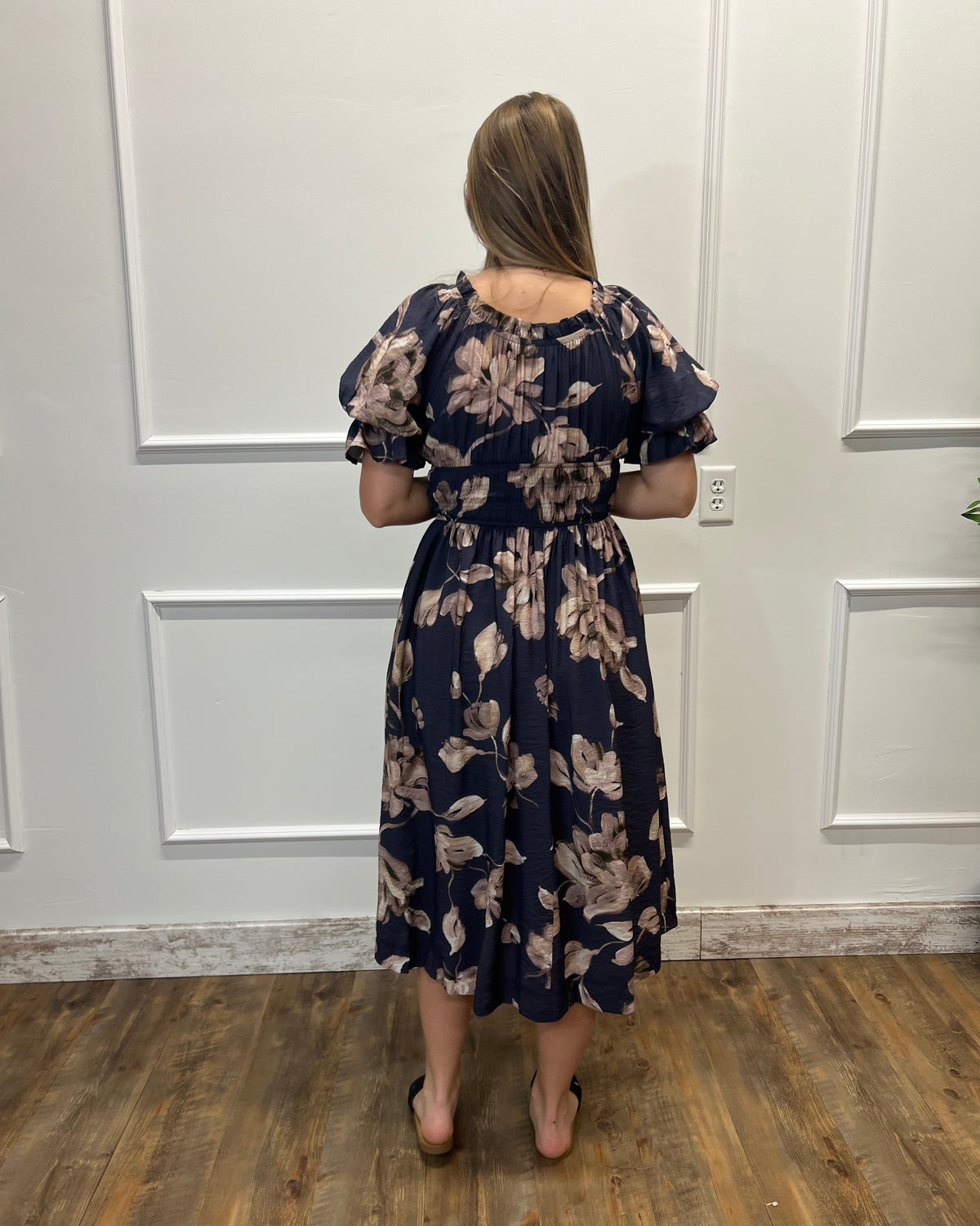 A woman viewed from behind wearing a navy blue midi dress with a beige floral pattern, puff sleeves, and a gathered waist, standing on a wooden floor against a white paneled wall.