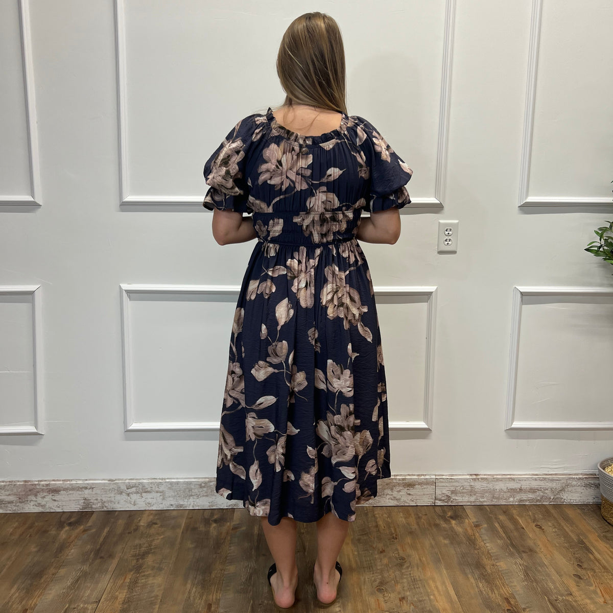 A woman viewed from behind wearing a navy blue midi dress with a beige floral pattern, standing on a wooden floor against a white paneled wall.