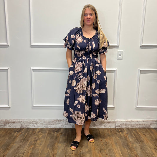 A woman stands indoors wearing a navy midi dress with a beige floral print, puff sleeves, and black slide sandals. She is positioned in front of a white paneled wall with a wood-look floor.