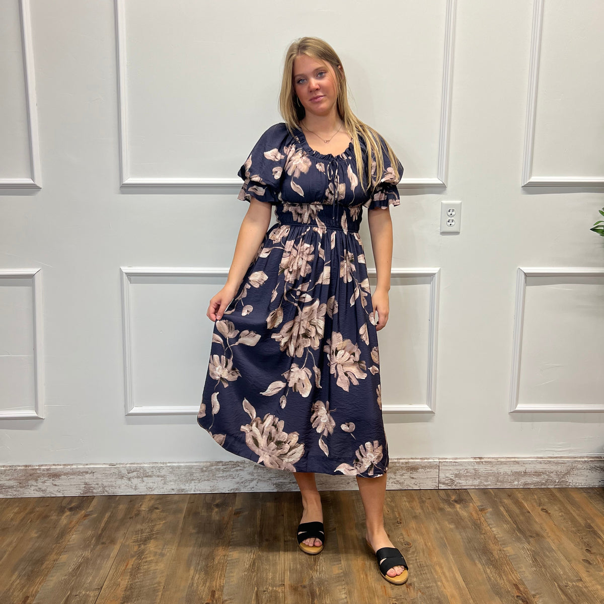 A woman modeling a navy blue midi dress with a beige floral pattern, standing in front of a white paneled wall on a wooden floor.