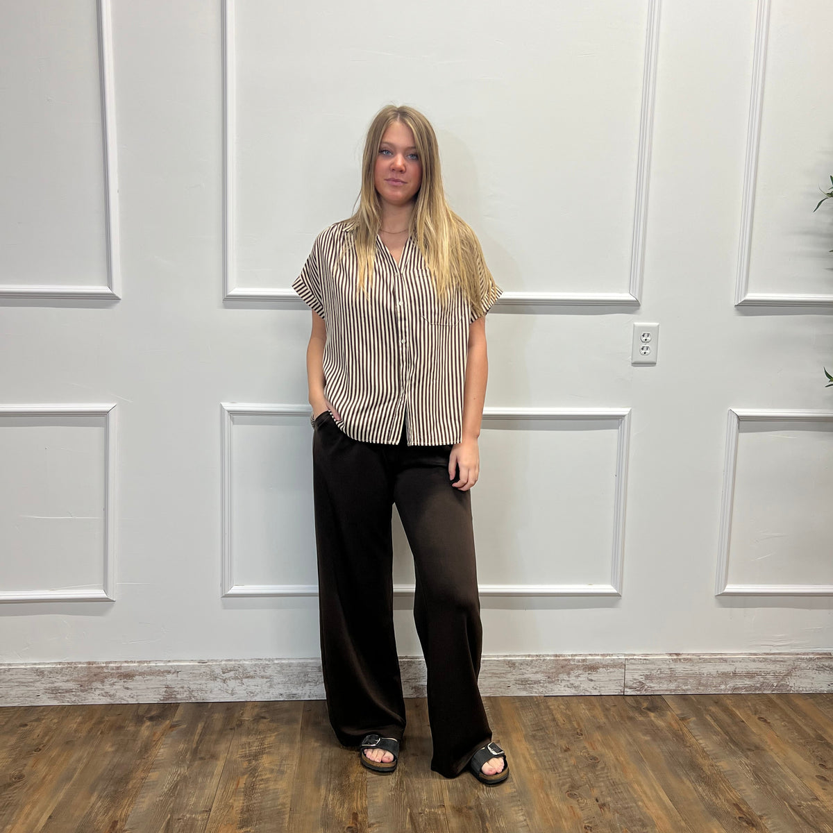 A woman stands in a room with white paneled walls and a wood floor, wearing a beige and black vertically striped short-sleeve button-up shirt, black wide-leg pants, and black slide sandals.