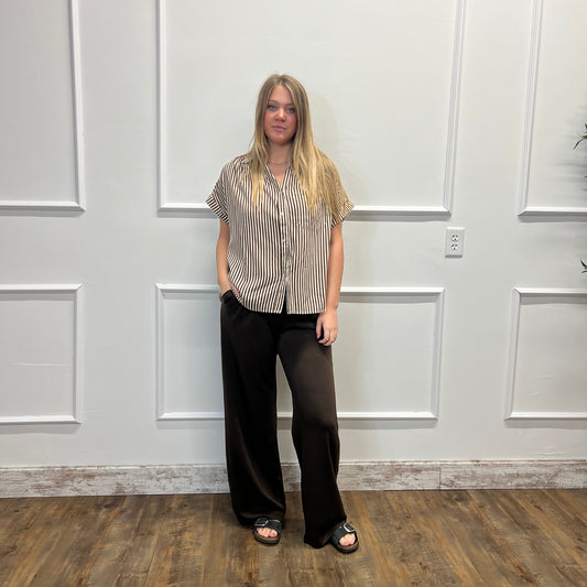 A woman stands in a room with white paneled walls and a wood floor, wearing a beige and black vertically striped short-sleeve button-up shirt, black wide-leg pants, and black slide sandals.