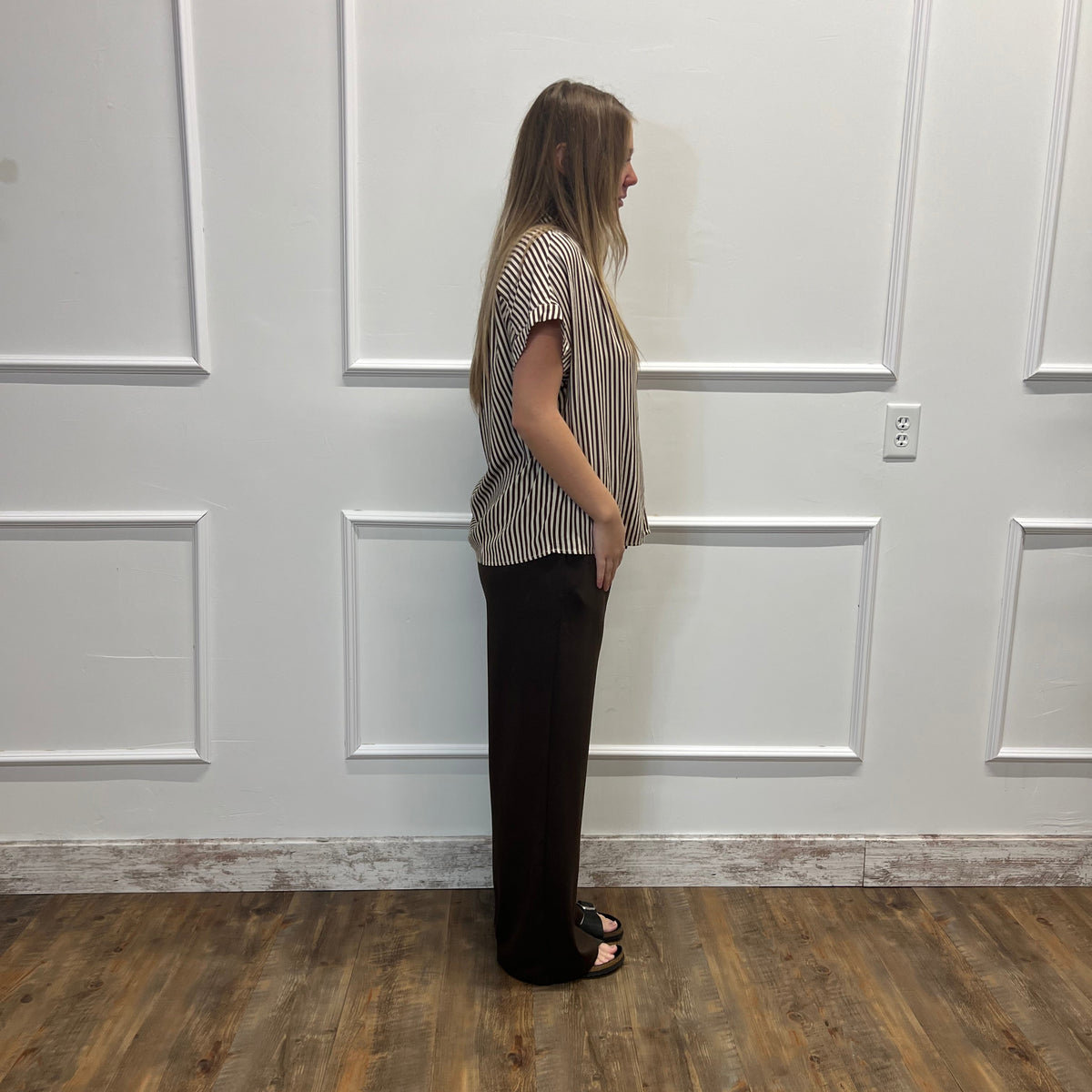A woman stands in profile wearing a short-sleeve striped top and dark wide-leg pants against a white paneled wall with wood-look flooring.