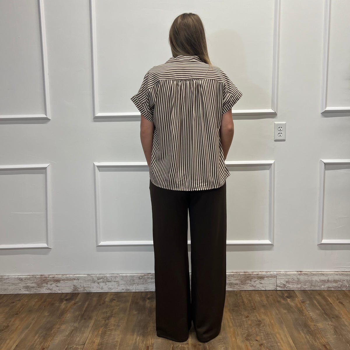 A woman viewed from behind wearing a short-sleeve button-up shirt with vertical brown and beige stripes, paired with dark brown wide-leg pants, standing in front of a white paneled wall on a wood floor.