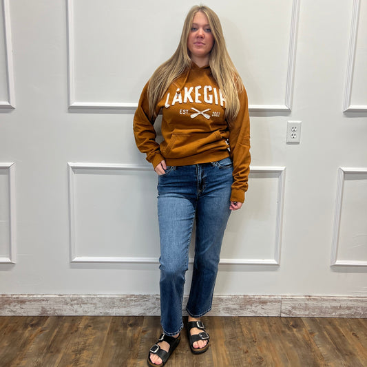 A woman stands against a white paneled wall wearing a brown sweatshirt with 'LAKEGEAR' printed in white, blue jeans, and black sandals.