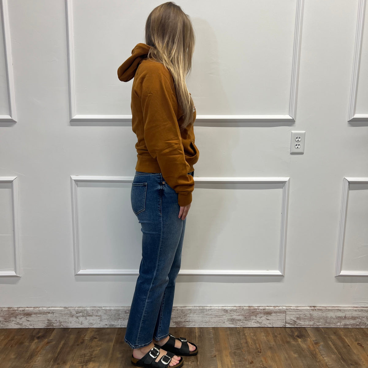 A woman stands in profile wearing a brown hooded sweatshirt, blue jeans, and black slide sandals, standing on a wood floor against a white paneled wall.