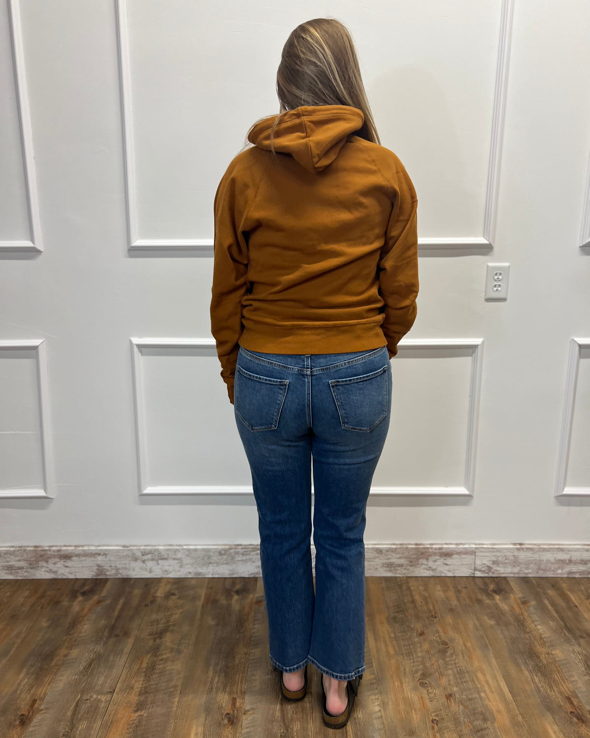 A woman viewed from behind wearing a rust-colored hooded sweatshirt and blue jeans, standing in front of a white paneled wall on a wood floor.
