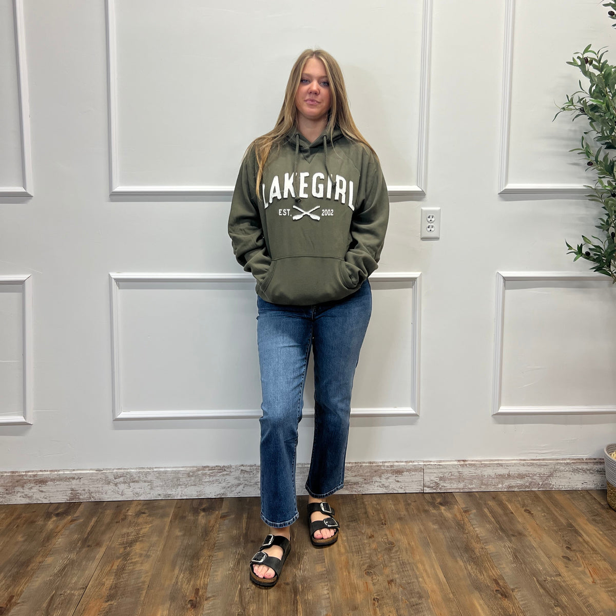 A woman stands in a room wearing an olive green hoodie with 'LAKE GIRL' printed in white across the chest, paired with blue jeans and black slide sandals.