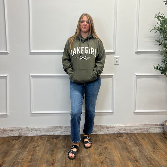A woman stands in a room wearing an olive green hoodie with 'LAKE GIRL' printed in white across the chest, paired with blue jeans and black slide sandals.