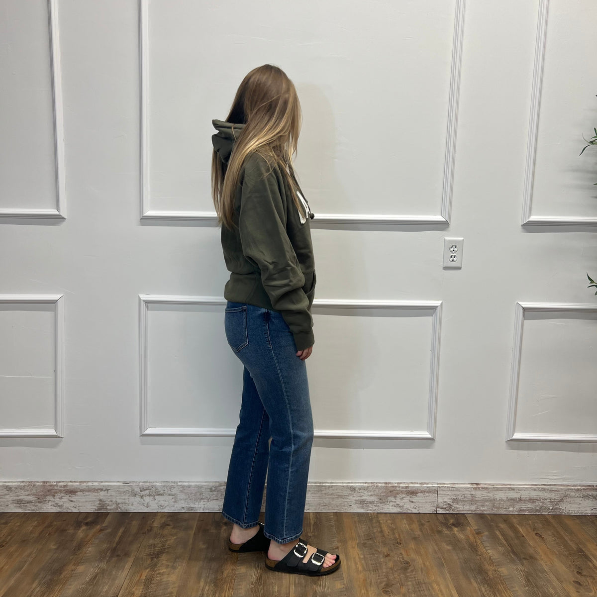 A woman stands in profile wearing an olive green hoodie, blue jeans, and black slide sandals, standing on a wood-look floor against a white paneled wall.