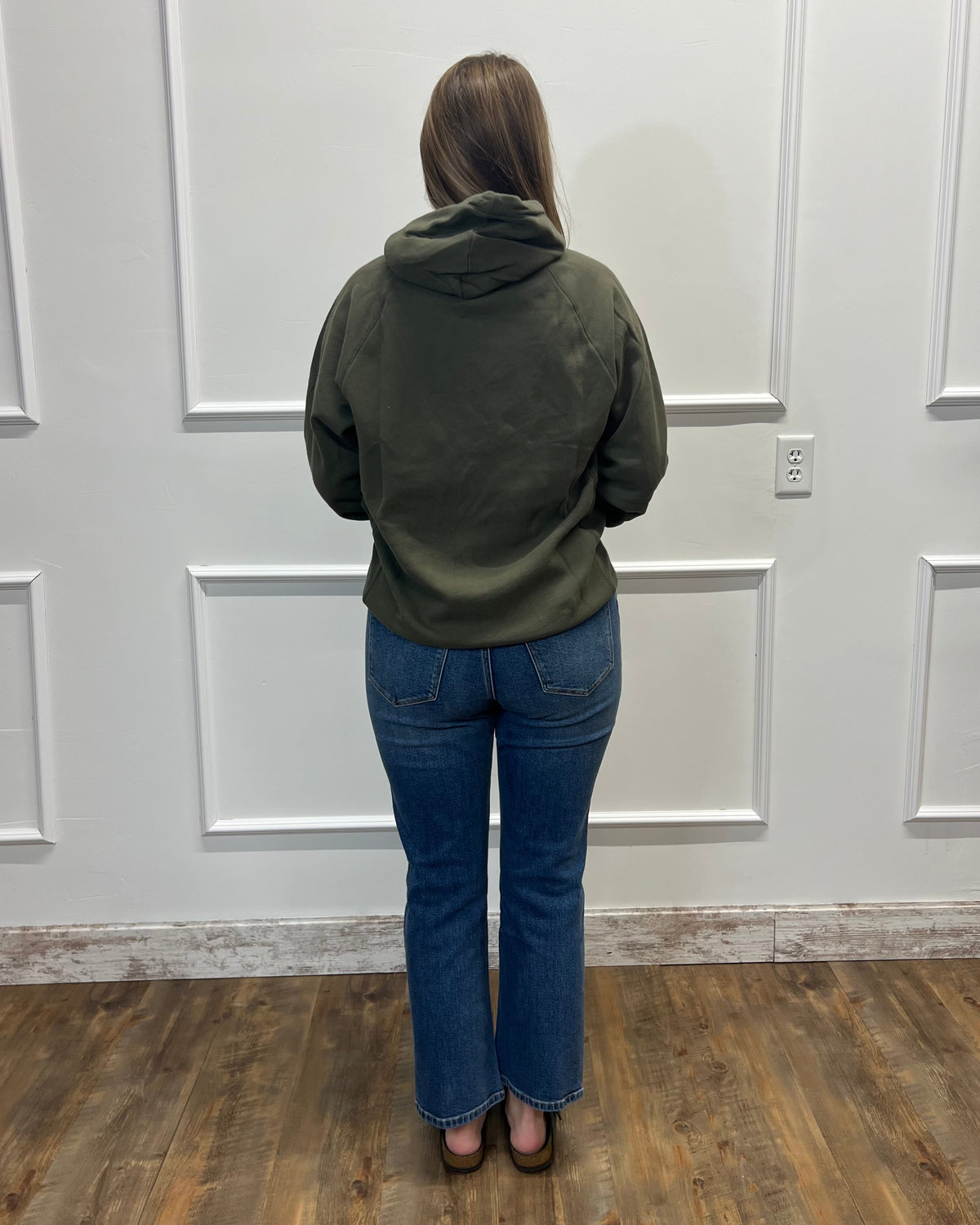 A woman viewed from behind wearing an olive green hooded sweatshirt and blue jeans, standing in front of a white paneled wall on a wood floor.