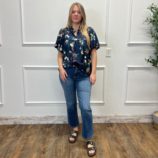 A woman stands indoors wearing a dark navy floral short-sleeve blouse with blue jeans and black sandals, posed against a white paneled wall with wood flooring.
