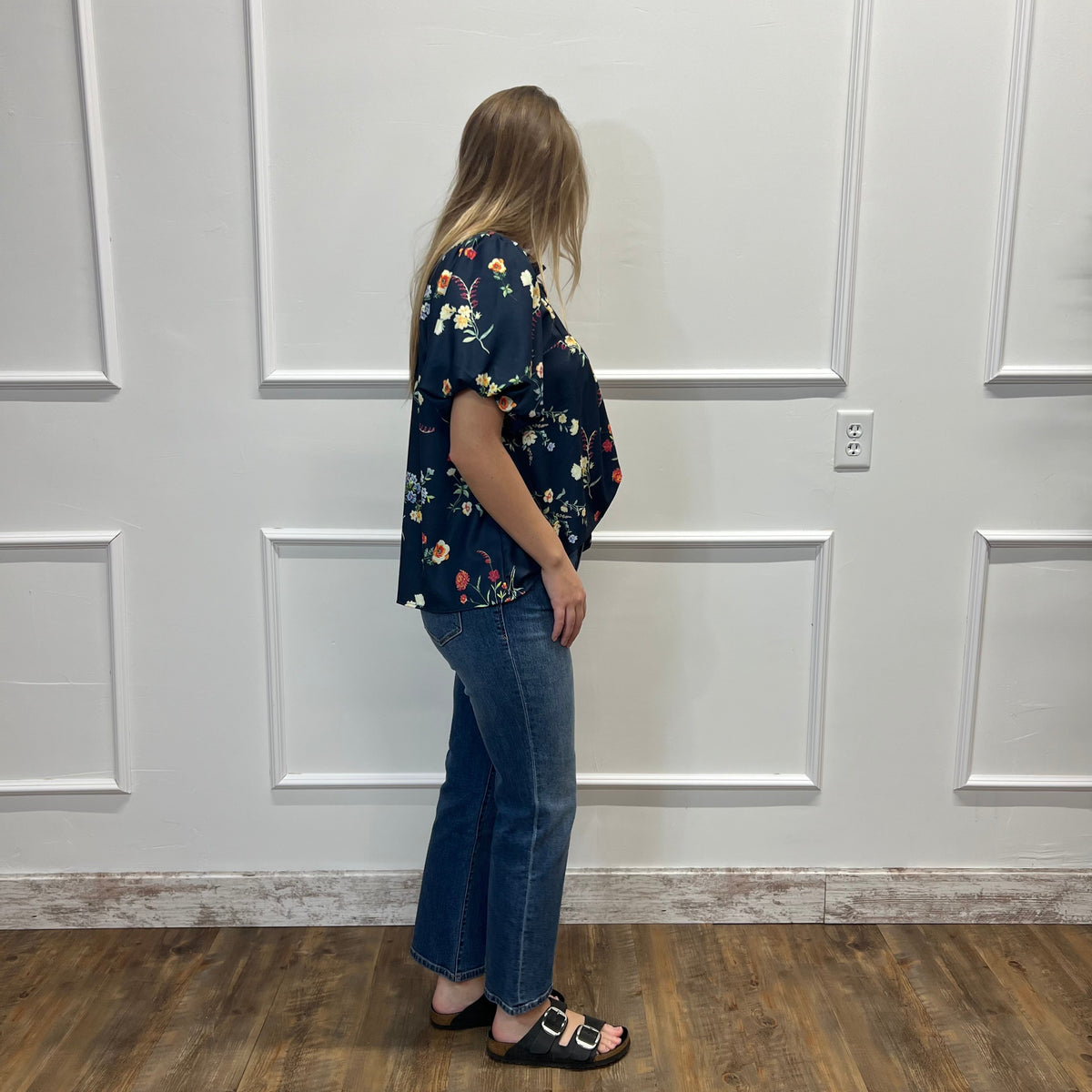 A woman stands in profile wearing a navy blue short-sleeve blouse with a colorful floral print, paired with blue jeans and black slide sandals, set against a white paneled wall and wood-look floor.