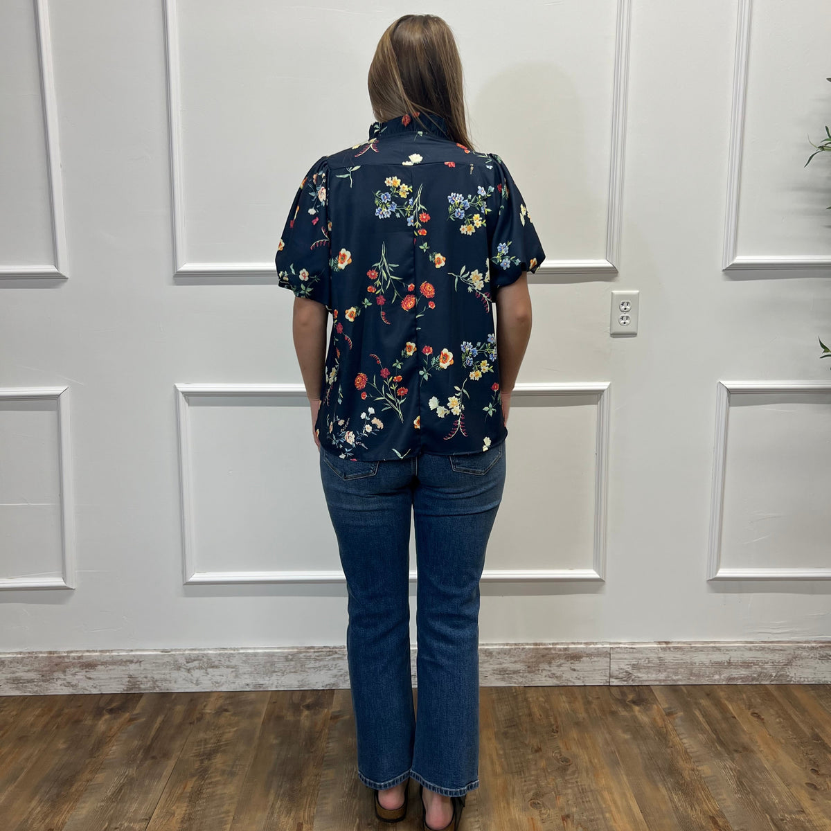 A woman viewed from behind wearing a navy blue short-sleeve button-up shirt with a colorful floral print and blue jeans, standing on a wood floor against a white paneled wall.
