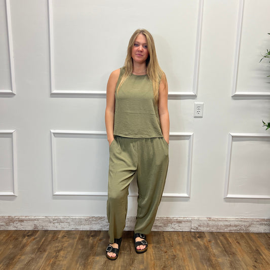 A woman stands indoors wearing a matching olive green sleeveless top and wide-leg pants set, paired with black sandals, in front of a white paneled wall on a wood floor.