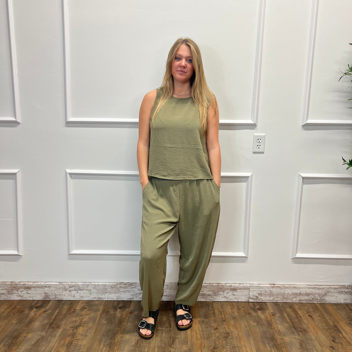 A woman stands indoors wearing a matching olive green sleeveless top and wide-leg pants set, paired with black sandals, in front of a white paneled wall on a wood floor.