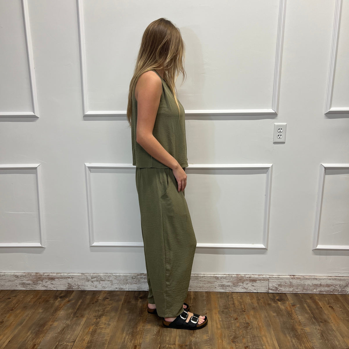 A woman stands in profile wearing a sleeveless olive green jumpsuit and black slide sandals, standing on a wood-look floor against a white paneled wall.