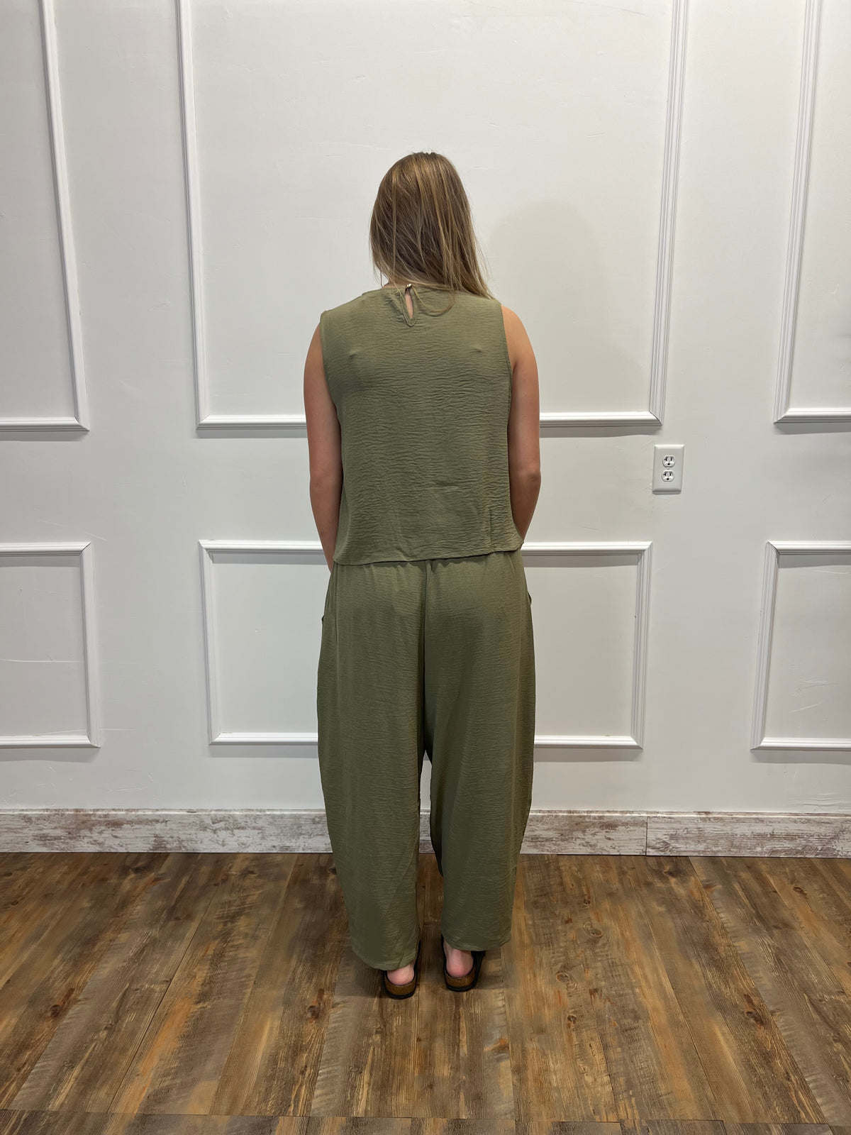 A woman stands with her back to the camera wearing an olive green sleeveless top and matching wide-leg pants set, standing on a wood floor against a white paneled wall.