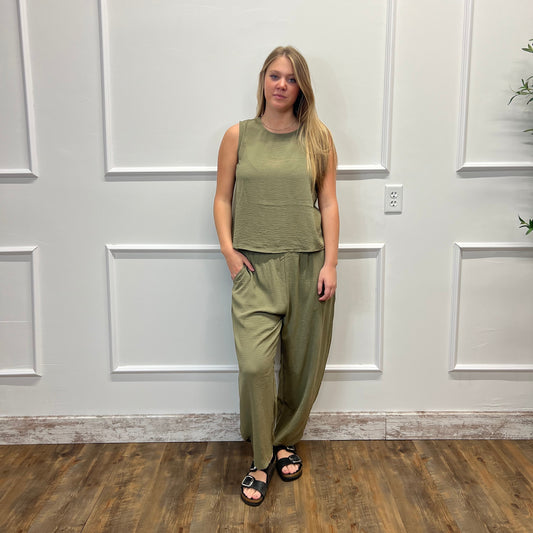 A woman stands indoors against a white paneled wall wearing an olive green sleeveless top and matching wide-leg pants, with black sandals, on a wood floor.