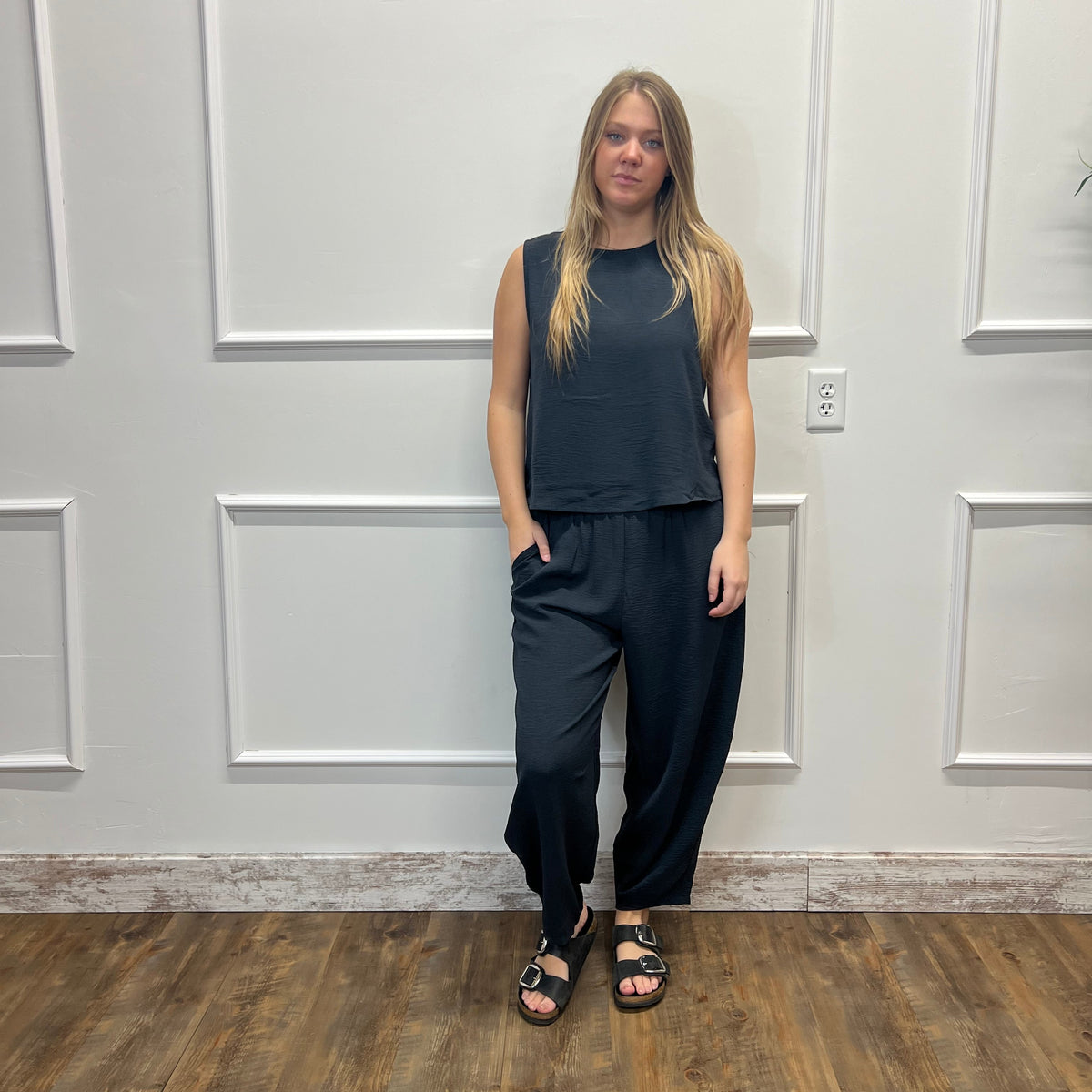 A woman stands in front of a white paneled wall on a wood floor, wearing a dark charcoal sleeveless top and matching dark wide-leg pants with black sandals.