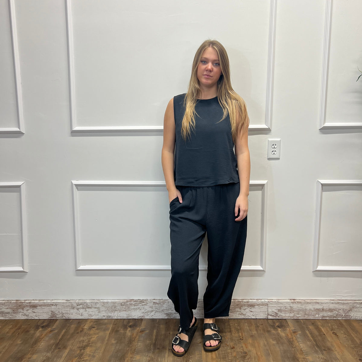 A woman stands in front of a white paneled wall on a wood floor, wearing a dark charcoal sleeveless top and matching dark wide-leg pants with black sandals.