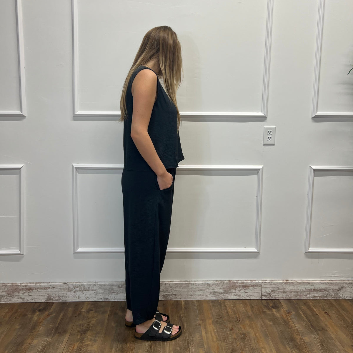 A woman stands in profile wearing a black sleeveless jumpsuit and black slide sandals, standing on a wood floor against a white paneled wall.