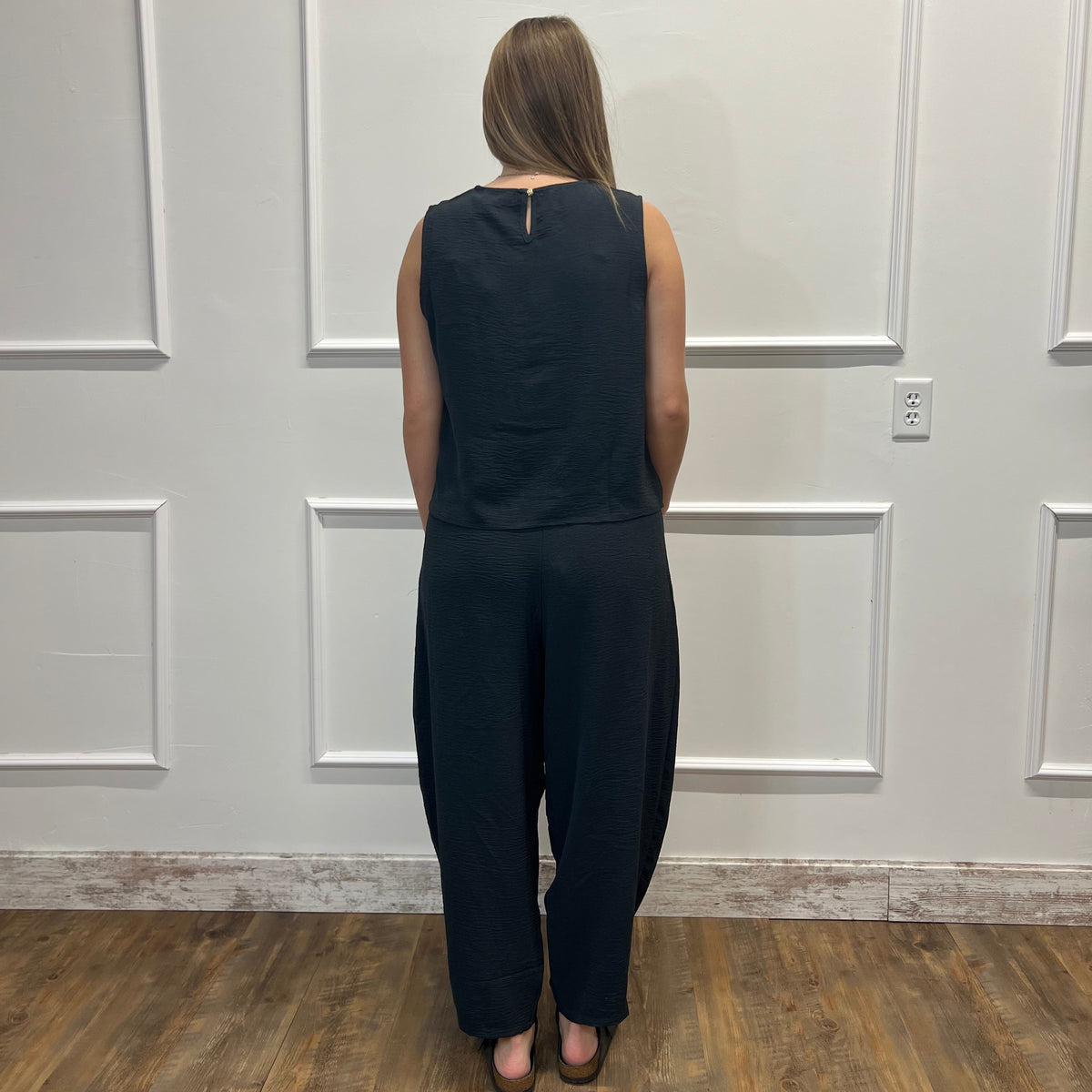 A woman viewed from behind wearing a sleeveless black jumpsuit with wide legs, standing on a wood floor against a white paneled wall.