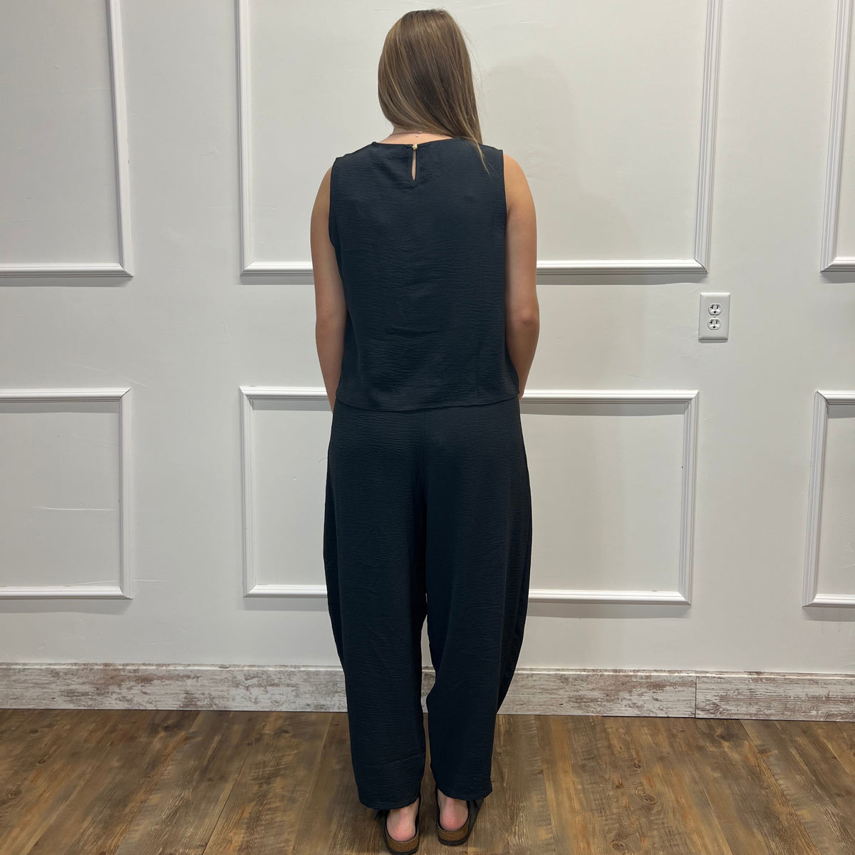 A woman viewed from behind wearing a sleeveless black jumpsuit with wide legs, standing on a wood floor against a white paneled wall.