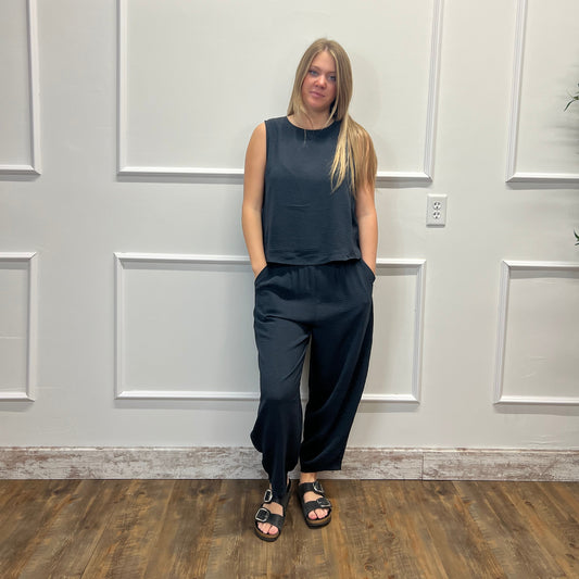 A woman stands against a white paneled wall wearing a dark slate blue sleeveless top and matching wide-leg pants, paired with black slide sandals.