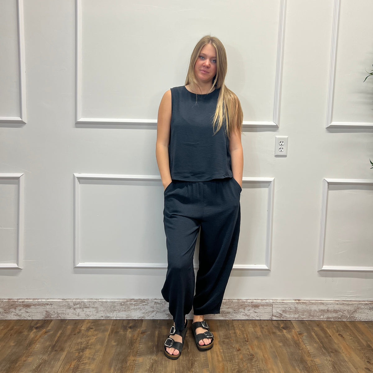A woman stands against a white paneled wall wearing a dark slate blue sleeveless top and matching wide-leg pants, paired with black slide sandals.