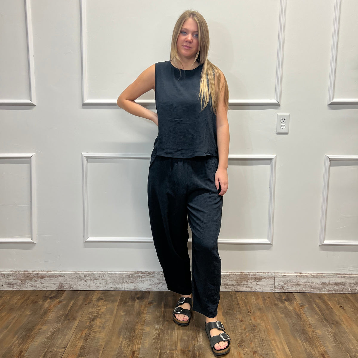 A woman stands in a room with white paneled walls and a wood floor, wearing a dark navy sleeveless top and matching dark navy wide-leg pants with black sandals.