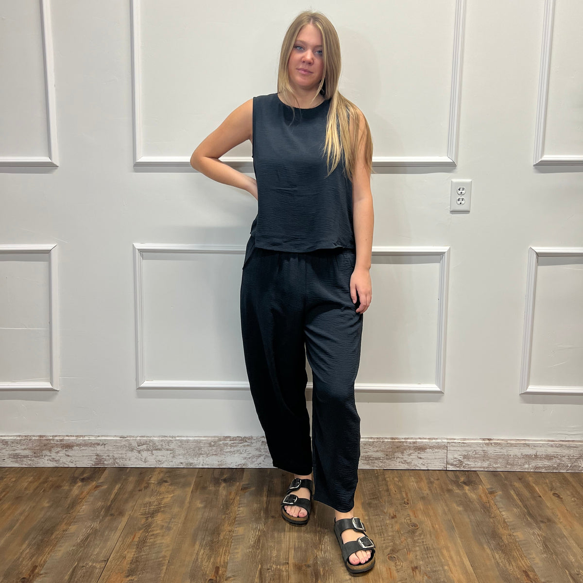 A woman stands in a room with white paneled walls and a wood floor, wearing a dark navy sleeveless top and matching dark navy wide-leg pants with black sandals.