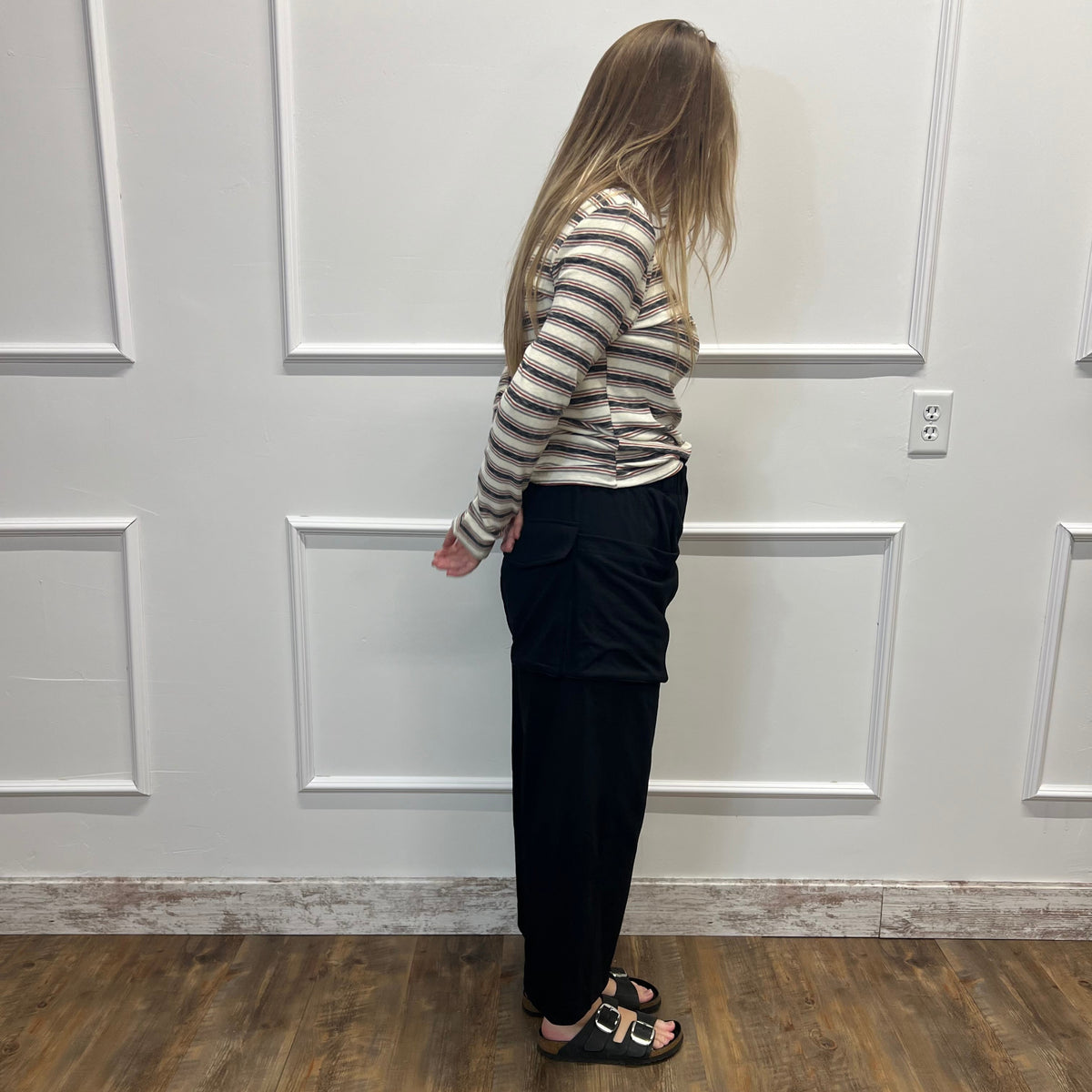 A woman stands in profile against a white paneled wall on a wood floor, wearing a beige and gray striped long-sleeve top, black pants, and black slide sandals.