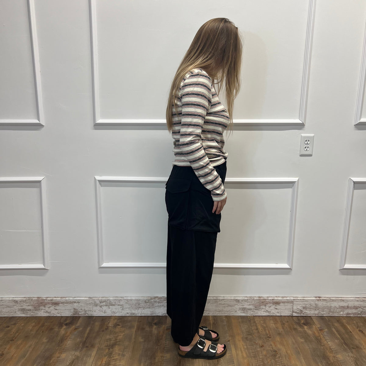 A woman stands in profile wearing a beige and gray striped long-sleeve top, black wide-leg pants, and black slide sandals with gold accents, set against a white paneled wall and wood-look floor.
