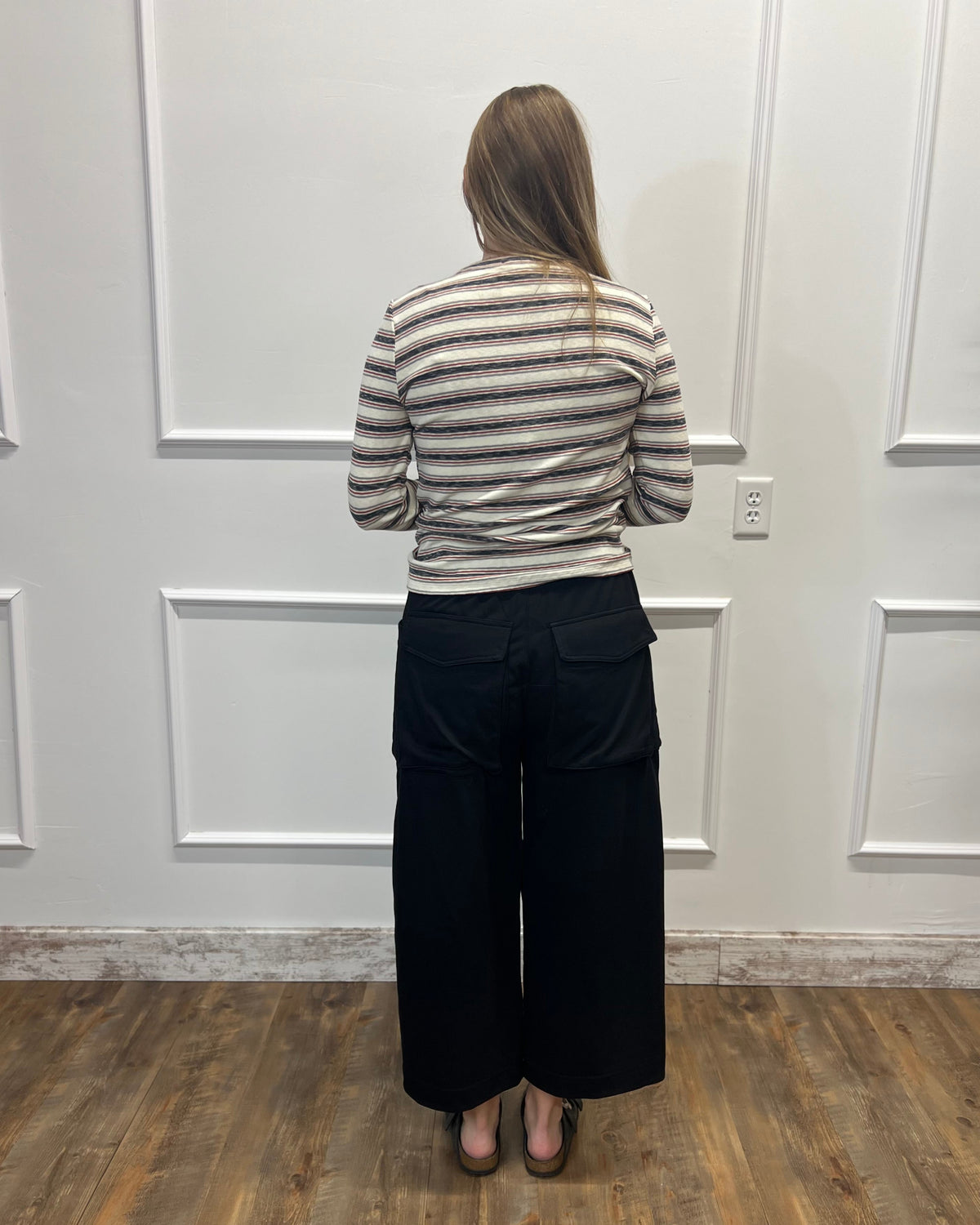 A woman viewed from behind wearing a beige and gray striped long-sleeve top and black wide-leg pants, standing on a wood floor against a white paneled wall.