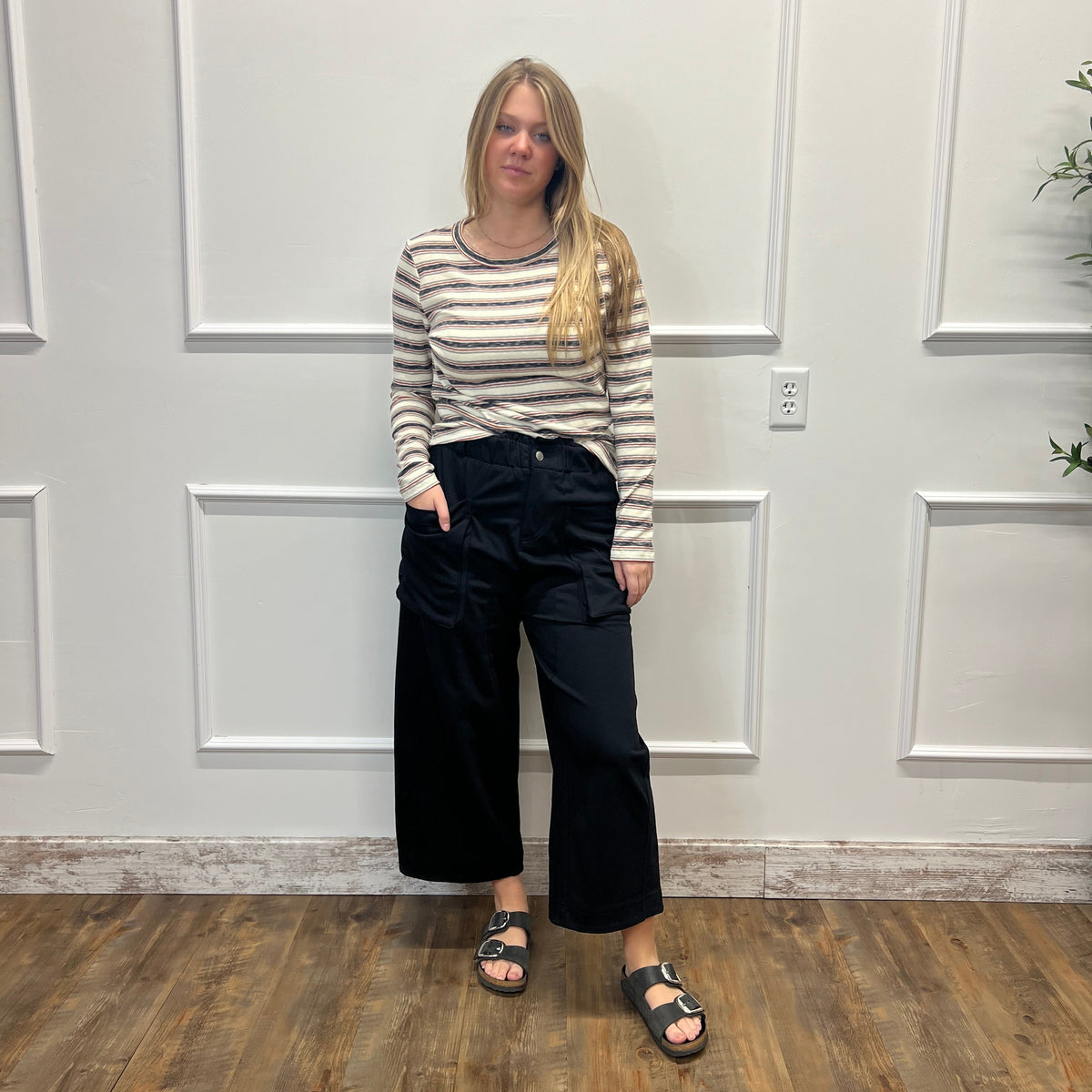 A woman stands in a room with white paneled walls and a wood floor, wearing a beige and white striped long-sleeve top, black cropped wide-leg pants, and black sandals.