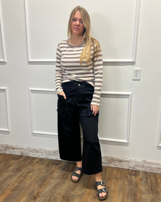 A woman stands in a room with white paneled walls and a wood floor, wearing a beige and gray striped long-sleeve top, black wide-leg cropped pants, and black sandals.