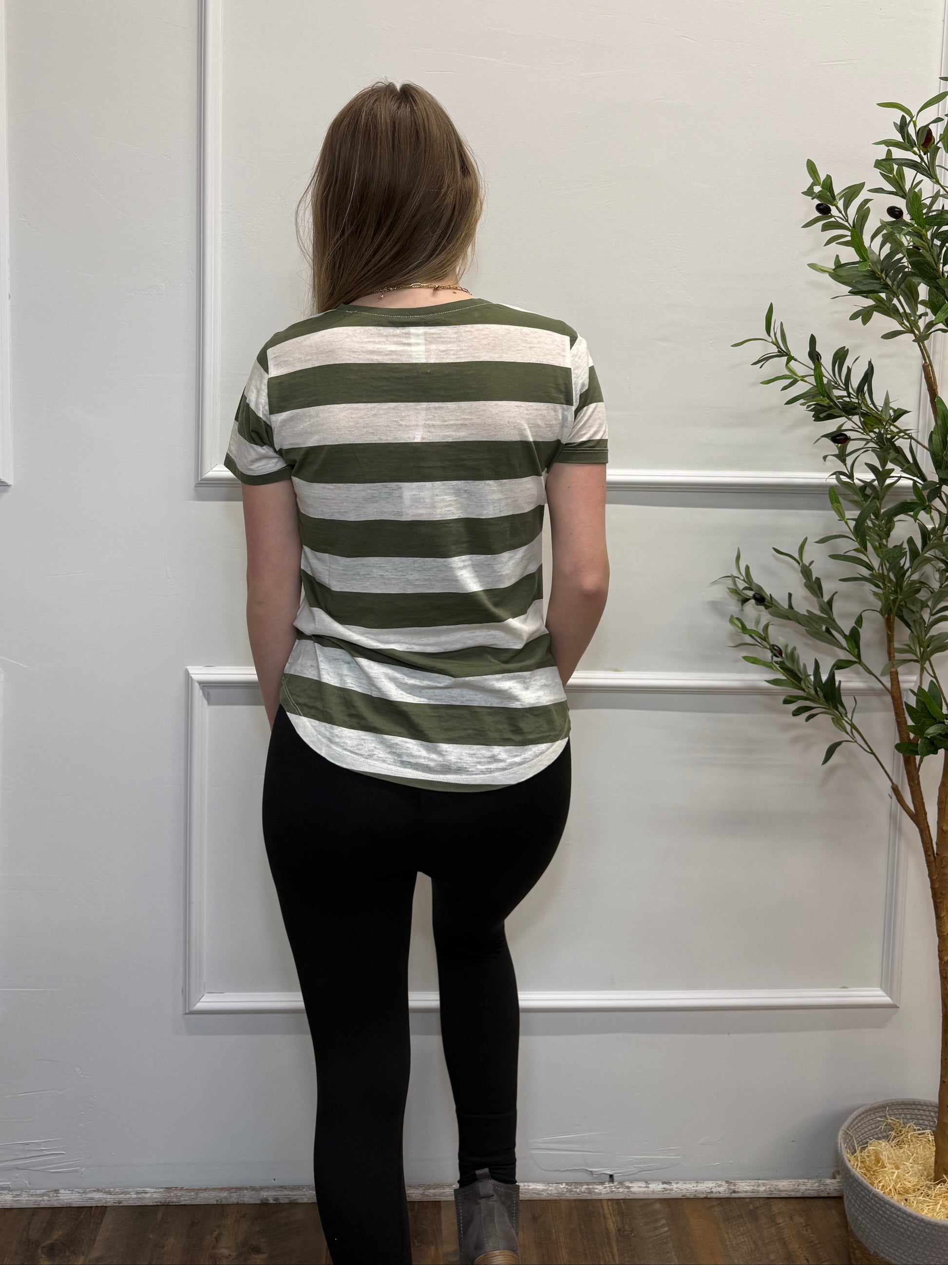 Person wearing a striped shirt and black pants standing in front of a white paneled wall with a plant.