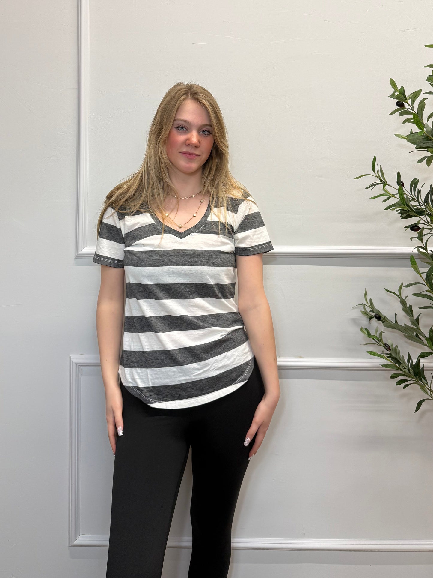 Woman wearing a black and white striped shirt and black pants standing against a white paneled wall with greenery.
