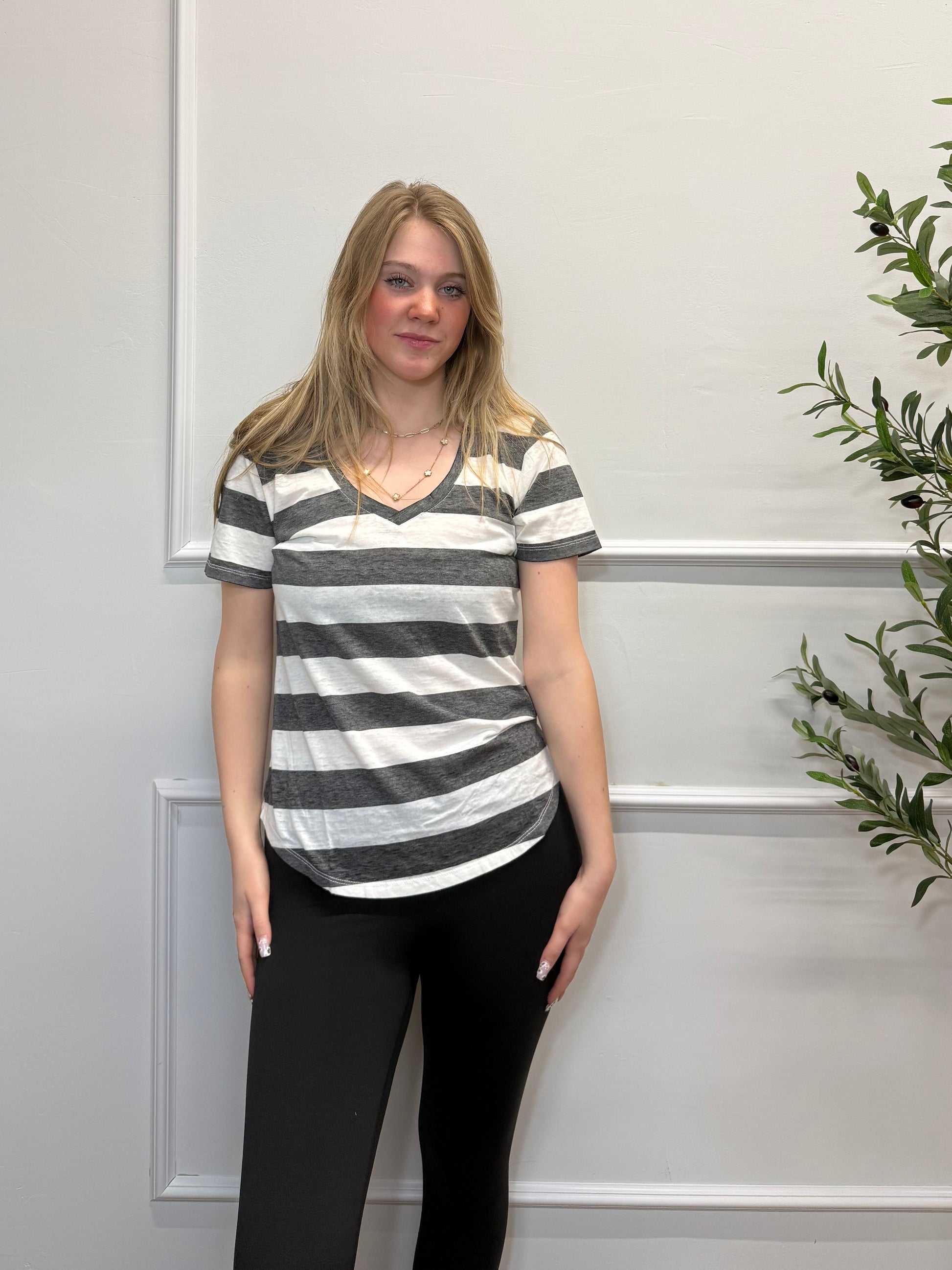 Woman wearing a black and white striped shirt and black pants standing against a white paneled wall with greenery.