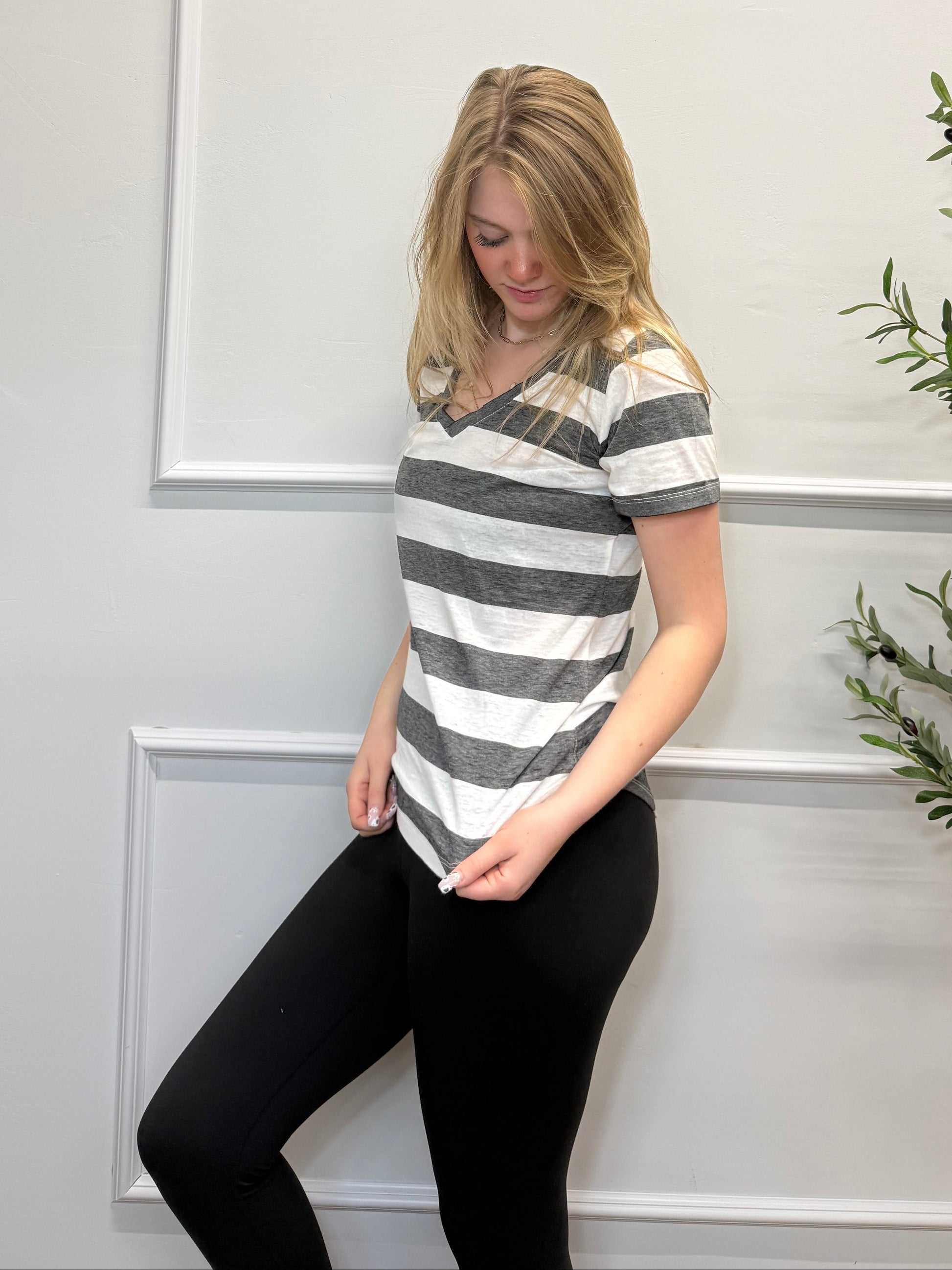 Woman in a striped shirt and black pants standing against a white wall with a plant.