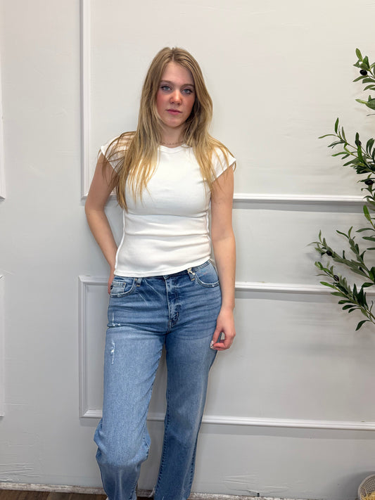 Woman wearing a white top and blue jeans standing in front of a white door with greenery.