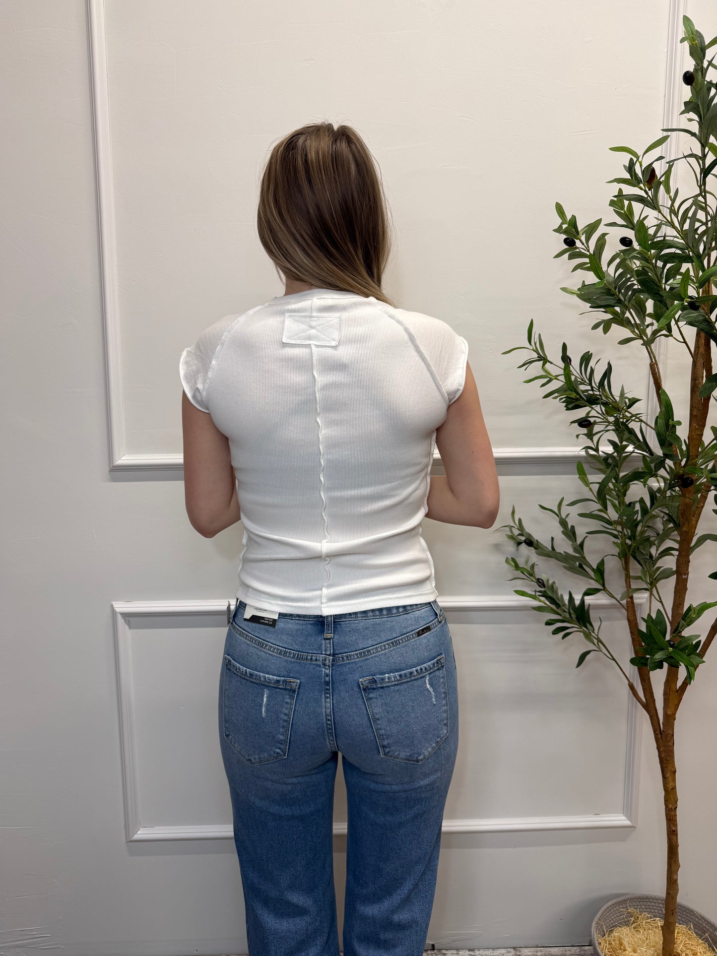 Person wearing a white tank top and blue jeans standing in front of a white door with a plant in the background.
