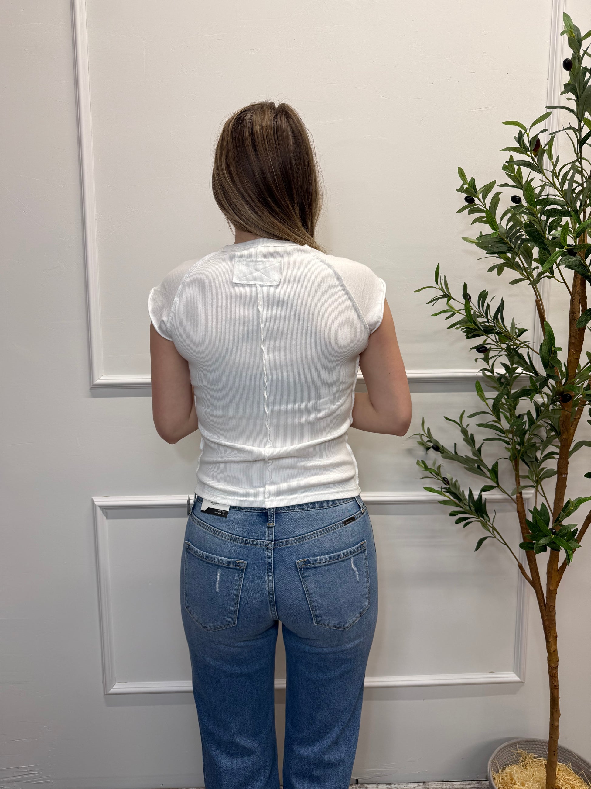 Person wearing a white tank top and blue jeans standing in front of a white door with a plant in the background.