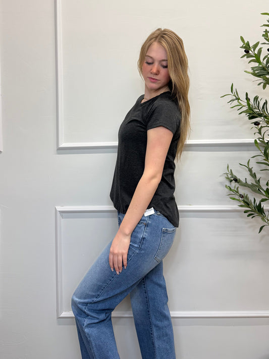 Person wearing a black shirt and blue jeans standing against a white paneled wall with greenery.