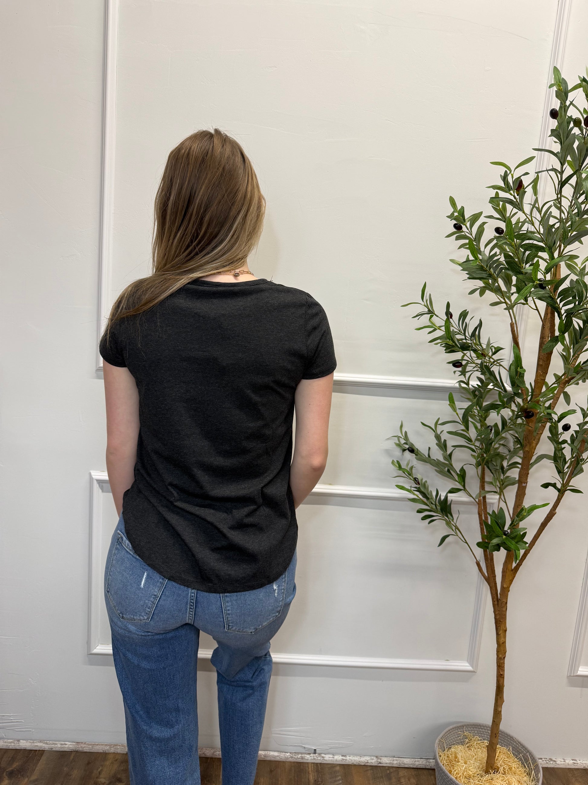 Person wearing a black shirt and blue jeans standing in front of a white door with a plant in the background.
